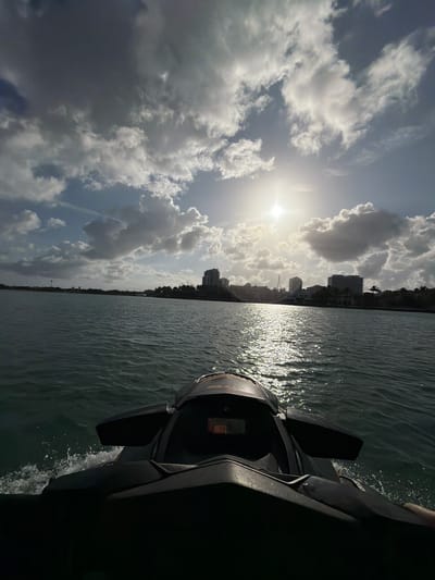 The image captures a scene from the water, likely taken from a watercraft, in Bal Harbour Village, United States of America. The sky is filled with a mix of clouds, some appearing dark and ominous, while others are bright white, reflecting the sunlight. The sun is positioned high in the sky, casting a glare. Below the sky, the water reflects the sunlight, creating a shimmering effect. The shoreline is visible in the distance, featuring buildings and vegetation. The watercraft is dark and partially obscures the view. The watercraft's wake is visible in the water.