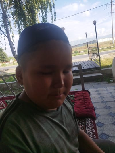 A young boy wearing a dark skullcap and a green t-shirt sits outdoors on a patterned mat. He is positioned on the right side of the frame, with his head tilted downwards and his eyes closed. Behind him, a wooden structure with a metal railing is visible, and further in the background, a road, a fence, and distant mountains can be seen under a partly cloudy sky. The setting appears to be an outdoor seating area, possibly a patio, in the city of Kyzyl-Kyya, Kyrgyzstan.