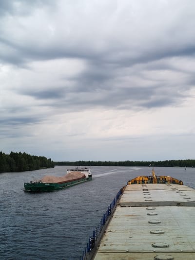 A wide, tranquil river flows under a cloudy, overcast sky, hinting at an approaching storm. The water is a deep, choppy blue, reflecting the muted tones of the sky. On the left bank of the river, a dense forest of green trees stretches as far as the eye can see, creating a natural, verdant border.
Two barges are prominent on the water. The barge closer to the left bank, a long, green vessel, is laden with a substantial load of light-colored gravel or sand, its cargo forming a rounded mound. The name "НЕВСКИЙ G" is visible on its side. This barge is moving away from the viewer, heading towards the distant treeline.
In the foreground, partially obscuring the view of the river and the other barge, is the deck of a much larger vessel. This barge is a pale, weathered gray, with numerous circular hatches visible across its surface, suggesting it is a cargo carrier designed for bulk goods. Its structure is robust and utilitarian, with a blue railing along the visible edge. The deck is clean but shows signs of wear and tear.
The overall scene is one of industrial transport set against a natural landscape. The weather is subdued, with the heavy clouds creating a dramatic and somewhat somber atmosphere. The time of day is likely afternoon or early evening, given the soft, diffused light. The presence of the barges and the cargo indicates ongoing commercial activity on the waterway, likely in the region of Novokemskii, Russia, given the location context. There are no people visible in the frame.