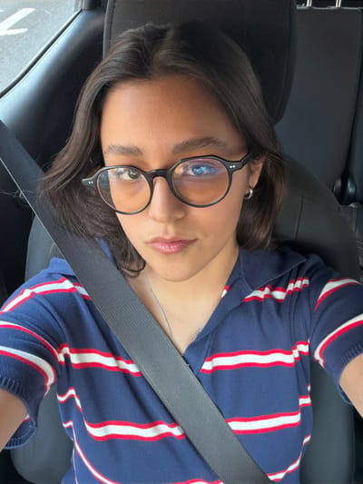 A young woman with dark hair and glasses is seated in a car. She wears a blue and white striped shirt and a seatbelt. The image is taken from above, showing her face and upper torso. The car's interior is dark, with a leather seat visible. She has a slight smile.