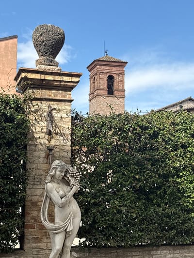 This image depicts an outdoor scene in Foligno, Italy, centered on a classical-style female statue. The statue, constructed from light-toned stone, features a partially draped female figure holding a cluster of grapes near her left shoulder and chest. Behind the statue, a brick pillar adorned with green climbing ivy is visible, topped by a dark, textured, urn-shaped stone finial. A dense wall of dark green foliage, possibly a hedge or ivy-covered wall, extends across the midground. In the background, a prominent brick bell tower with arched belfry openings and a tiled roof with a finial and weather vane is visible against a clear blue sky with sparse white clouds. A section of a reddish-brown building facade is present in the far left background. No human individuals are present, and the scene is static under daylight conditions.