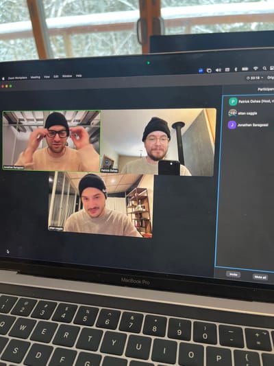 A MacBook Pro screen displays a Zoom meeting with three participants. Patrick Oshea (Host), Eitan Caggia, and Jonathan Saragossi. Each is visible in their individual video feeds, showing their faces. The keyboard of the MacBook Pro is also visible to the right of the screen. The Zoom interface includes options like "Mute all" and "Invite." Text on the top menu bar includes: Zoom Workplace, Meeting, Edit, Window, and Help. The system status indicates a time of 3:18.