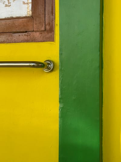 The image is a close-up shot of a wall with a horizontal chrome grab bar mounted on the yellow section. The wall is painted in two distinct colors, yellow and green, separated by a vertical line. A wooden window frame with a glass pane is visible in the upper left corner, partially obscuring the wall. The grab bar is positioned centrally in the yellow area, extending from the left edge towards the green section. The scene appears to be an interior or exterior wall, possibly in a public space given the presence of the grab bar. The location is identified as Pasekaran, Indonesia.