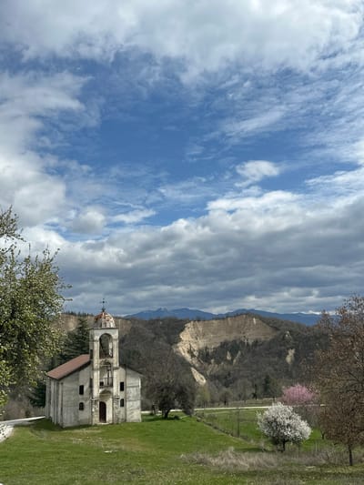 An external view captures a scene in Rozhen, Bulgaria, featuring a weathered church or chapel positioned on a verdant grassy knoll. The church, constructed from light-colored masonry, possesses a prominent bell tower with an arched opening and a reddish, domed roof topped with a cross. Dark, rectangular windows punctuate its sides, and a dark wooden door marks the entrance. A narrow paved path ascends towards the structure from the left.
The immediate foreground consists of green grass interspersed with patches of drier vegetation. Beyond the church, the landscape transitions into hills characterized by extensive, light-tan geological formations resembling eroded cliffs or pyramids, indicative of the Rozhen Pyramids. Denser, dark green forest covers other sections of these hills. Further in the midground, a solitary white-blossomed tree stands out on the right, with another pink-blossomed tree nearby. Trees with nascent green foliage are visible on the far left.
In the distant background, mountains, some capped with snow, rise beneath a sky that exhibits a mixture of clear blue patches and expansive, white to grey stratocumulus clouds. No individuals or discernible actions are present within the frame.