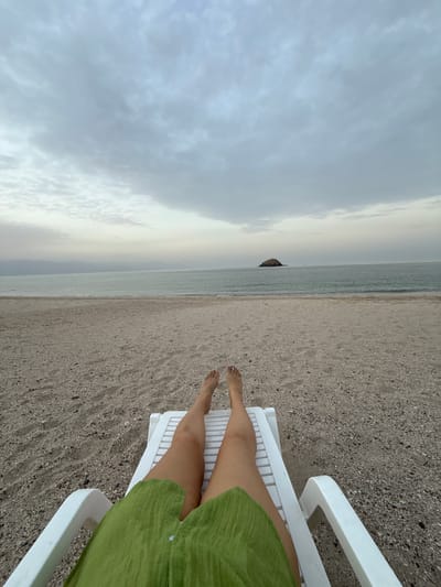The image depicts a person relaxing on a white plastic beach chair at a beach in Dhadna, United Arab Emirates. The viewpoint is from above, looking down the length of the person's legs towards the horizon. The person is wearing a green garment. The beach consists of light gray sand, and the water is a muted blue-gray color. The sky is overcast with thick, gray clouds. A small, rocky island is visible in the distance, just off the shoreline.