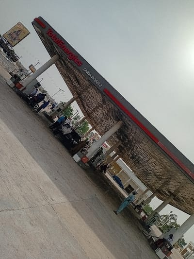 The image displays a TotalEnergies gas station identified as "ZARIA ROAD 2," located in Kariya, Nigeria. The scene is illuminated by bright natural light, with sun glare visible in the upper right. A large, sloped grey and red canopy, bearing "TotalEnergies" branding, covers the station's forecourt and is supported by several white pillars. The underside of the canopy exhibits a textured pattern and some discoloration.
Beneath the canopy, multiple grey fuel pumps are positioned. Approximately ten individuals are present across the forecourt. One person in a light blue uniform stands in the lower right foreground, facing away from the viewer. Another individual in a white uniform is visible further back near a pump on the right. A cluster of several people is gathered on the left side, near what appears to be a motorcycle or small vehicle. People are standing or moving, indicating general activity at the station.
The forecourt is paved with concrete, showing visible cracks and wear. In the background, beyond the main pumping area, additional light-colored buildings, green vegetation, and a large billboard displaying an advertisement are visible. A line of vehicles, including what appear to be trucks, is present on an adjacent road in the far left background.