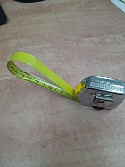 A close-up, eye-level shot shows a yellow metal tape measure lying on a light-colored wooden surface. The tape measure is extended and curved, with the metal casing of the tape measure visible on the right side of the frame. The tape itself has black markings and numbers, with the numbers 14, 15, 16, 17, 18, 19, 20, 21, 22, and 23 clearly visible. The wooden surface appears to be a tabletop with a visible grain pattern. The scene is well-lit, with soft shadows. There are no people or other objects clearly visible in the frame. The context of the location in the city of Shomron Regional Council, Palestinian Territories is not discernible from the image itself.