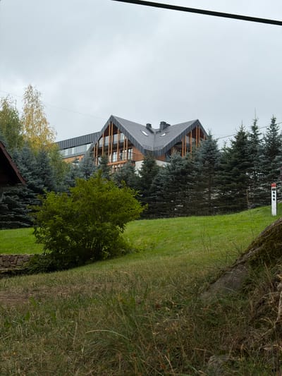 A modern, multi-story building with a dark, angular roof and wooden accents is visible behind a line of evergreen trees. A patch of green lawn is in the foreground, with a large, leafy bush in the center. The sky is overcast. The building appears to be a hotel or resort, possibly located in the mountain town of Świeradów-Zdrój, Poland, known for its spa resorts and scenic beauty. A white post with red accents is visible to the right of the image. The overall scene suggests a tranquil, natural setting.