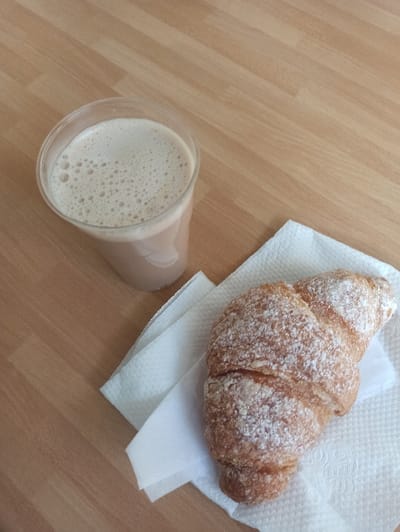 A clear plastic cup filled with a light brown, foamy beverage is positioned on a light-colored wooden surface. To the lower right of the cup, a golden-brown croissant, dusted with white powdered sugar, rests on a folded white paper napkin. The wooden surface exhibits a diagonal grain pattern. The scene depicts an unattended breakfast or snack arrangement in an indoor setting within San Marzano sul Sarno, Italy.