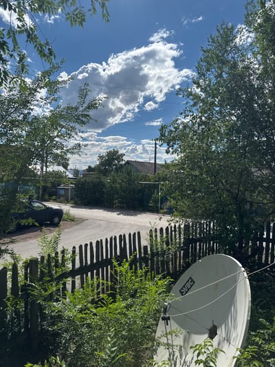 The image depicts a residential scene in Abay, Kazakhstan, on a bright, sunny day with scattered white clouds against a blue sky. The perspective is from a slightly elevated viewpoint, looking down a paved residential road lined with trees and a wooden fence.
Prominently featured in the upper right is a white satellite dish, mounted on a pole and surrounded by lush green foliage. The dish has a visible logo that appears to be "ST*S" or similar. A dark-colored car is parked on the side of the road, partially obscured by trees. In the background, several houses with pitched roofs and brick structures are visible, suggesting a rural or suburban setting. Power lines crisscross the sky, indicating the presence of infrastructure.
The lighting suggests it is daytime, likely midday given the strong sunlight and distinct shadows cast by the fence and trees onto the road. The overall mood is tranquil and ordinary, capturing a slice of everyday life in this location. No people are visible in the frame.