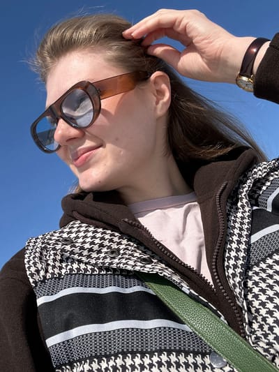 This is a close-up, low-angle portrait of a young woman standing outdoors under a clear blue sky in Новое Шигалеево, Russia. The subject, a woman with light brown hair, is positioned with her head slightly tilted and her gaze directed towards the viewer's right. She is smiling faintly. Her right hand is raised, with fingers gently touching the hair near her right temple. She wears oversized, brown-framed sunglasses with reflective blue lenses. Her attire consists of a light pink or lilac-colored crew-neck shirt, layered under a dark brown, possibly fleece, zip-up hoodie or jacket. Over these, she wears a vest or jacket featuring a prominent black and white houndstooth pattern, interspersed with horizontal black and light grey striped sections. Several dark buttons or snaps are visible on the upper left front of this patterned garment. A dark green, textured bag strap runs diagonally across her chest. A gold-toned watch with a dark brown strap is visible on her right wrist. The background is an unobstructed bright blue sky, indicating clear weather.