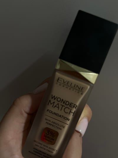A person is holding a bottle of Eveline Wonder Match Foundation in their hand. The bottle is made of glass with a black cap and a gold accent. The label on the bottle is beige and has black text. The text on the bottle says "EVELINE COSMETICS WONDER MATCH FOUNDATION SKIN ABSOLUTE PERFECTION 3% SERUM complex SECOND SKIN EFFECT 24H SATIN FINISH HYALURONIC ACID THERMAL WATER MICROBIOME PR". The background is dark and out of focus. The lighting is dim. The image appears to be taken indoors.