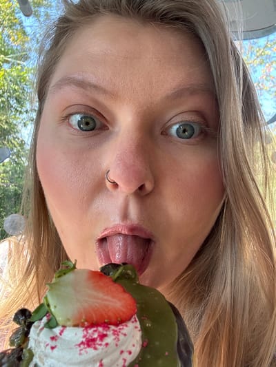 A close-up, eye-level image captures an individual, likely female, presenting her face directly to the camera while interacting with a dessert. Her wide-open, light-colored eyes (blue/green) gaze forward, and her mouth is open with her tongue extended downwards, appearing to lick or taste the food item. A silver ring pierces her left nostril. Light brown or blonde hair frames the sides of her face. Below her mouth, a dessert is partially visible. It consists of a pastry, likely a donut, topped with a generous portion of white whipped cream, a prominent sliced red strawberry, several dark berries (possibly blueberries), and a vivid green glaze (resembling matcha) with pink sprinkles. The background is blurred but shows green foliage and bright daylight, suggesting an outdoor or glass-enclosed natural environment. No specific identifying markers for the location in San Phra Net, Thailand, are visible within the image.