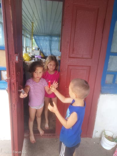 Three children are standing in a doorway. On the left, a young girl with curly dark hair wears a pink striped t-shirt and white underwear. She holds a silver door handle. To her right, a younger girl with blonde hair wears a pink t-shirt and also appears to be wearing underwear. Her right hand is extended, holding a red object. To the right of the doorway, a young boy in a blue sleeveless shirt and black athletic shorts with white stripes is seen from the side, reaching towards the girls. The background shows an interior space with a blue curtain with white stars, and various items on a table including a yellow bag and a purple bowl. Outside the doorway, a white bucket with green leaves and some other organic material is visible. The overall environment appears to be rustic or rural, possibly a porch or entryway to a home. The image is watermarked with "nubia Focus 5G" in the bottom left corner.