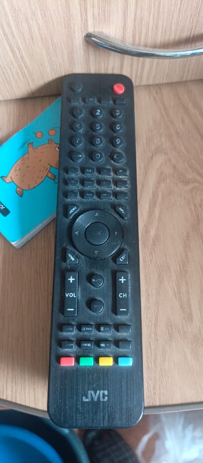 The image is a close-up, eye-level shot of a black JVC television remote control resting on a light-brown wooden surface, likely a table or shelf. The remote is positioned vertically, with its top slightly to the left and its bottom to the right. A portion of a blue notepad with a drawing of a brown bear is visible on the left side of the frame, partially obscuring the remote. The wooden surface has a visible grain pattern. The remote control itself is rectangular with rounded corners and has numerous buttons. At the top, there is a red power button. Below that, there are rows of numbered buttons (1-9), along with other function buttons. A circular navigation pad with an "OK" button in the center is located in the middle section of the remote. Below the navigation pad, there are buttons labeled "HOME," "VOL +," "VOL -," "CH +," and "CH -." Further down are colored buttons: red, green, yellow, and blue. The "JVC" logo is visible towards the bottom of the remote. The scene is well-lit, with no shadows obscuring the details of the remote. The overall environment appears to be indoors, with the wooden surface and the presence of electronic devices suggesting a domestic or office setting. There are no people visible in the image, and no actions are taking place. The image is static. The location is not identifiable as Sokol, Russia.