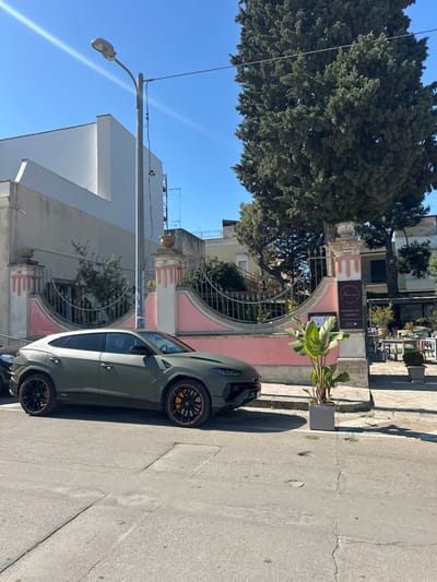 The image captures a street scene in Nardò, Italy, featuring a light olive green Lamborghini Urus parked along the side of the road. The foreground shows the car parked next to a concrete sidewalk with cracks and imperfections. Behind the car, there is a light pink building with a decorative gate, partially obscured by a large leafy tree. A sign indicates a restaurant or bistro. Across the street, a white building stands under a clear blue sky, and a lamppost is visible. The scene appears to be captured during daylight hours, with shadows cast by the objects.
