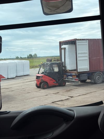 In Zarech'e, Russia, a red Linde forklift is actively engaged in moving large white bales, which are wrapped in plastic. The forklift is positioned on a concrete surface, with several other identical bales stacked to its left. Behind the forklift and bales, a shipping container is open, and the forklift appears to be in the process of loading or unloading contents into it. The container is mounted on a dark-colored truck. The environment is an outdoor industrial or storage area, with a flat landscape and a cloudy sky. The image is taken from the interior of a vehicle, possibly a truck cab, looking out through the windshield.