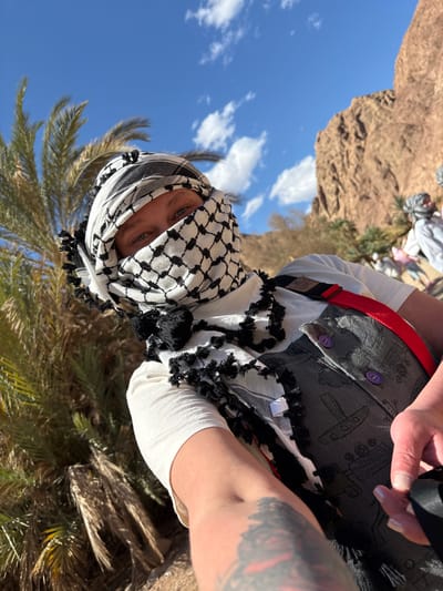 A low-angle, medium shot depicts a person wearing a traditional black and white checkered keffiyeh headdress with fringe. The person is outdoors, and part of a palm tree is visible near the bottom of the frame, suggesting an oasis or desert environment. The sky is blue with a few scattered clouds. The person is wearing a white long-sleeved shirt, a patterned gray vest with purple buttons, and a red strap. Part of a tattoo is visible on the person's arm. In the background, other people are present amidst tan terrain. Based on the mention of Dahab, Egypt, the location likely represents a desert landscape or an oasis setting in that region.