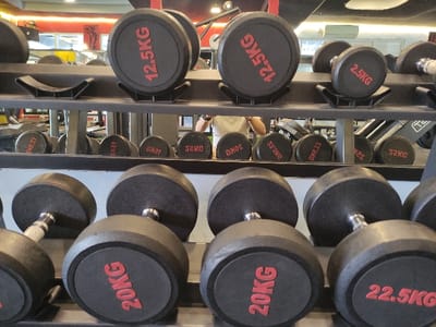 The image depicts an indoor gym setting. There are no identifiable features within the image to confirm a specific location within the city of Staufenberg, Germany.
The primary subjects are multiple rows of dumbbells arranged on a black metal storage rack. In the immediate foreground, a bottom tier of the rack displays several black rubber-coated dumbbells with silver or chrome handles. Visible weight denominations in red lettering on black circular ends include "20KG" and "22.5KG". Above this, a second tier holds additional dumbbells, with "12.5KG" and "2.5KG" clearly visible on their ends.
Behind the foreground dumbbell rack, a large reflective surface, likely a mirror, extends across the scene. This mirror reflects a third row of dumbbells, showing denominations such as "30KG", "32.5KG", "35KG", "42.5KG", and "52KG" (some text appears reversed due to reflection). The reflection also reveals other gym equipment, including a dark-colored fitness machine, possibly a treadmill or elliptical, further in the background. A partial view of an individual wearing a light-colored shirt is visible in the mirror's reflection, standing among the gym equipment. The environment includes a dark ceiling with visible lighting fixtures and a red accent wall visible in the upper left background reflection. No specific actions or interactions beyond the static arrangement of equipment are observable.
