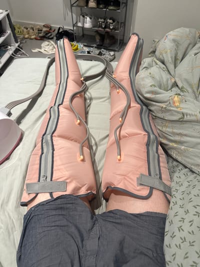 The image shows a person lying in bed, wearing compression boots on both legs. The boots are pink with gray zippers and trim, and are connected by gray tubing to a white device located to the left of the frame. The person is wearing dark gray shorts. The bed is covered with a light gray sheet and a comforter with a green botanical print. In the background, a metal shoe rack holds several pairs of shoes, and a white shelf unit is partially visible. The overall scene suggests a domestic setting, likely a bedroom. There is no information in the image to indicate the location is Vilanova i la Geltrú, Spain.