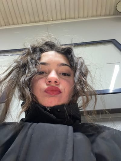 A woman in a dark winter coat poses for a selfie. Her wavy brown hair is slightly disheveled. She has light skin, dark eyes, and is wearing red lipstick, puckering her lips slightly. The background includes a white wall, a window frame, and a hint of what appears to be a radiator. The photo appears to be taken indoors and rotated.