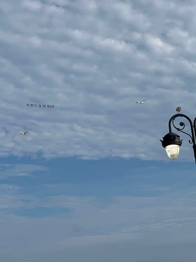 A banner pulled by a small airplane reads "NO BUTTS ON THE BEACH!" The banner is in the sky above the ocean. A second airplane is visible in the distance. A lamppost with a bird on top is in the foreground. The sky is cloudy. The location is Ocean City, United States of America.