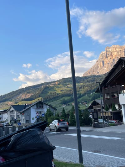 The image captures a picturesque mountain village scene, likely in La Villa, Italy. The orientation suggests the photo was taken in portrait mode and then rotated. The foreground is dominated by a dark, possibly metal, dumpster overflowing with a large black plastic bag. A grey pole runs vertically through the image, obscuring parts of the scene.
Beyond the dumpster, a street with clear white lane markings curves into the distance. A white car is visible on the road. The buildings lining the street are typical of a European alpine village, with dark wood accents and balconies. Several traffic signs are present, including a blue directional sign with an arrow and a circular sign with a green cross.
The mid-ground reveals the lush green slopes of a mountain, dotted with trees and small clusters of houses. The mountain itself rises dramatically in the background, its rocky, sun-drenched face contrasting with the verdant slopes. The sky is a vibrant blue, with scattered white clouds adding depth and texture.
The lighting suggests it is daytime, possibly late afternoon, given the warm light on the mountain face. The weather appears clear and pleasant. The overall mood is serene and idyllic, showcasing the natural beauty of the Dolomites juxtaposed with the charm of a small mountain community. No discernible text is visible on the buildings or signs, except for license plates on the vehicles.