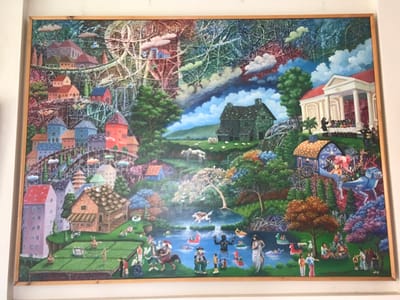 This image depicts a large, framed, vibrant painting, reportedly located in Kota Batu, Indonesia. The painting presents a complex, fantastical, and densely populated landscape.
**Scene Description:**
The composition is divided into multiple interconnected zones. On the left, an urbanized hillside features numerous multi-colored, multi-story houses clustered together, connected by elevated walkways and bridges. A large Ferris wheel and a roller coaster-like structure are prominent amidst a background of abstract, tangled lines and colored clouds in red, blue, and green hues. Cable car pods are visible along elevated tracks. Below these, a large, flat green field hosts various activities, including figures engaged in what appears to be farming, a game resembling volleyball, and social gatherings. A brown building with striped awnings stands adjacent to this field.
The upper right portion of the painting features a classical white building with columns and a red roof, where a crowd of figures dressed in black robes and white collars stands before a central orator. Further left on a hill, a dark green, multi-gabled house is surrounded by green pastures with white sheep.
The central and lower right sections of the painting depict a diverse natural and recreational environment. A body of water, possibly a lake or river, contains numerous individuals swimming or on colorful floats, including a black, ape-like figure. On the banks and in the foreground, diverse people are depicted: a figure in white drapery stands by the water, groups socialize or picnic on the grass, and individuals on horseback are present. A highly ornate, blue and gold gypsy-style carriage or float is depicted on an elevated track, seemingly pulled by a blue, stylized mythical creature, with a figure possessing pink hair inside. The surrounding terrain is lush with varied trees and colorful foliage, creating a vibrant, dreamlike atmosphere that merges urban, rural, and fantastical elements.