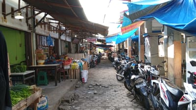 This is a medium-shot, eye-level, outdoor photograph of a market street in Puloara, Indonesia. The street is paved with hexagonal stones and is lined with stalls and shops on both sides. A variety of motorcycles are parked along the right side of the street, creating a narrow passage. Several blue and multi-colored tarps are stretched overhead, providing shade. On the left side of the street, there are stalls selling produce, including bundles of green onions and leafy vegetables in a wooden crate. A person is seated at a table inside one of the shops, and another person is partially visible in the foreground on the far left. The overall impression is of a busy, traditional market environment.