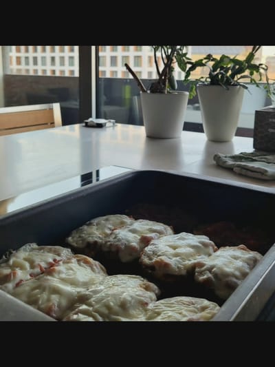 In a bright, indoor setting with a large window overlooking an urban landscape of multi-story buildings, a rectangular metal baking pan holds several round food items topped with melted cheese. The food appears to be some kind of patties or cutlets with a red sauce underneath the cheese. The pan is placed on a white table, which also features a small black object with white elements, possibly a napkin dispenser. To the left of the table, a portion of a wooden chair with a metal frame is visible. In the background, two white ceramic pots with green plants sit on the table, near the window. The overall scene suggests a meal being prepared or served indoors. The context of Bielawska Wieś, Poland, is external information and cannot be inferred from the image itself.