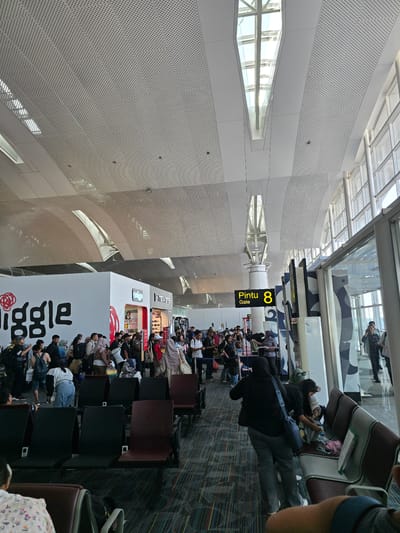 The image depicts a busy airport departure lounge. A large group of people, appearing to be passengers, are standing and milling about, many with luggage. In the background, a white structure with the word "Jiggle" in large black lettering is visible, possibly a store or kiosk. Further back, a sign with "Pintu 8 Gate" indicates a specific boarding gate. The environment is modern, with a high, perforated ceiling and large windows allowing natural light to enter. Rows of seating are present, some occupied and some empty. A few individuals are seen walking towards or away from the gate area. The scene suggests an active transit point, with passengers awaiting their flights. The location is identified as Ramunia Dua, Indonesia.