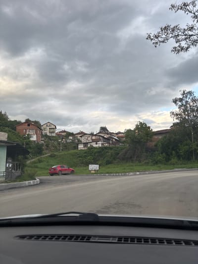 A medium shot taken from inside a vehicle shows a street with a row of houses on a grassy hillside, under an overcast sky. A red car is parked on the roadside. The location is likely in Baylovo, Bulgaria, as specified.
