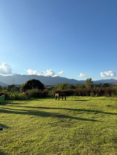 The image depicts a panoramic view of a natural landscape in Ban Mae Ping, Thailand, under a clear blue sky with scattered white cumulus clouds. The foreground consists of a well-maintained, sunlit green lawn, displaying long shadows indicative of late afternoon lighting. A small, dark brown, ornate wooden table or bench, with four legs and a flat surface, is positioned centrally in the mid-ground of the grass. To the far left, partially visible, is a vibrant green object, possibly a tent or a large piece of fabric. The mid-ground beyond the lawn features dense, wild vegetation, including various green shrubs and trees, some with reddish-brown foliage. Faintly visible on the left horizon, above the treeline, are power lines or transmission towers. The background is dominated by a majestic, distant mountain range, partially obscured by cloud cover. No individuals or active subjects are present within the frame; the scene is devoid of human activity.