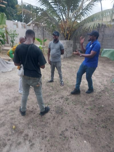 The image portrays an outdoor scene in Mwandege, Tanzania, featuring four male subjects on a dirt and sparse vegetation ground. In the foreground, a male stands with his back to the camera, wearing a black short-sleeved shirt, light grey trousers, and black sneakers, with a white bag slung over his left shoulder. Partially visible behind him and slightly to his right, another individual wears a multi-colored, patterned garment. To the left of center, a male stands facing right, dressed in a grey horizontally striped polo shirt, grey trousers, dark sandals, and a dark baseball cap. To the right of center, a male stands facing left, wearing a blue polo shirt, blue jeans, dark shoes, and a dark baseball cap, with both hands raised and open in a gesturing motion, suggesting active communication. A small, yellow, spherical object rests on the ground in the lower-left foreground. The background features a large palm tree bearing yellow coconuts, a stack of grey concrete masonry units, and a light-colored concrete block wall along the rear perimeter. A section of a light-colored building with a window is visible on the far left. The sky is overcast.