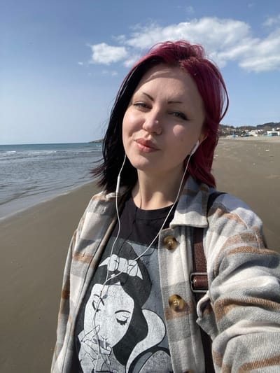 An individual is depicted in a close-up, selfie-style photograph taken on a sandy beach in Ulcinj, Montenegro. The subject is a female, appearing to be in her mid-20s to early 30s, with a two-toned hairstyle featuring black hair on the left side and magenta-red hair on the right. She is looking directly at the camera with a slight smile and pursed lips, wearing white wired in-ear headphones. Her attire consists of a black t-shirt featuring a white graphic print of a woman, possibly Snow White, with closed eyes and a hand near her mouth, layered under an open light gray and brown plaid shirt or jacket with brown buttons. A brown leather bag strap is visible over her right shoulder.

The background reveals a wide, calm body of water, likely the Adriatic Sea, on the left, with gentle waves reaching the shoreline. The sandy beach extends into the distance on the right, where distant low-rise buildings and a treeline are visible along the coastline, indicative of a coastal settlement. The sky is clear blue with scattered white cumulus clouds. The scene is illuminated by natural sunlight, suggesting a bright day.