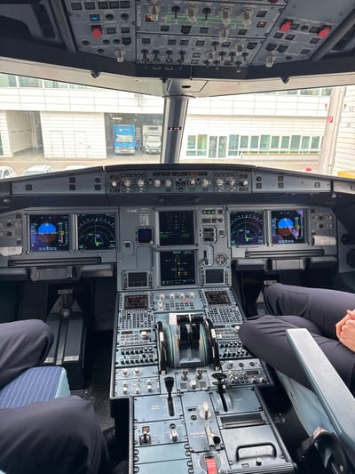 The image depicts the interior of an Airbus A320-214 cockpit, specifically the aircraft with registration D-AIND, stationary at a gate at Munich Airport (MUC), located in Oberding, Germany.
**Scene Description:**
The perspective is from within the cockpit, facing forward through the windshield. Two individuals, presumably pilots, are visible in the pilot and co-pilot seats, primarily showing their legs clad in dark uniform trousers. The pilot in the right seat has their hands clasped in their lap.
The cockpit instrumentation is fully visible and illuminated, including the overhead panel, glare shield, primary flight displays (PFDs), navigation displays (NDs), Electronic Centralized Aircraft Monitor (ECAM) displays, and the central pedestal. The central pedestal features twin thrust levers in an idle position, various control panels, and communication/navigation radios. The multi-function displays show data, including what appears to be a weather radar and flight path information.
Through the cockpit windshield, an outdoor airport environment is visible. The aircraft is positioned next to a terminal building, with an airbridge structure partially visible on the right. In the middle distance on the apron, two trucks are parked; one is blue, and the other is white. The background consists of a modern, light-colored airport building with horizontal architectural elements and numerous windows. The outdoor lighting suggests an overcast or diffused daylight condition.