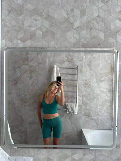 A young woman with blonde hair is taking a selfie in a bathroom mirror. She is wearing a teal sports bra and matching bike shorts. Behind her, a silver heated towel rack holds two white towels. The bathroom walls are tiled with a marble-like pattern, and the floor has a geometric tile design. The image appears to be taken indoors, likely in a residential bathroom. There is no information provided about the location in Berd', Russia.