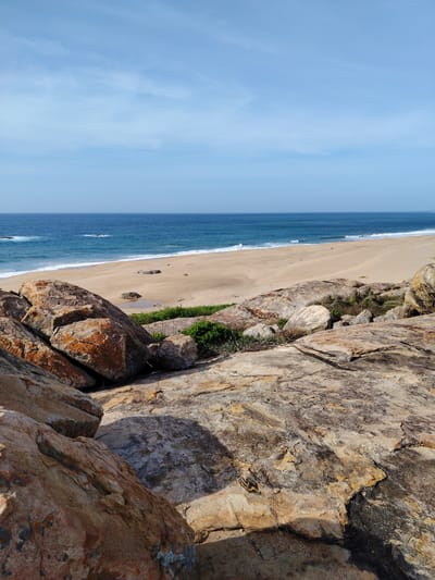 The image depicts a coastal landscape on a sunny day. In the foreground, large, weathered rocks with hues of orange, brown, and grey dominate the scene. These rocks are unevenly shaped and scattered across a sloping terrain. Between the rocks, patches of dry grass and small green shrubs are visible.

The middle ground features a wide expanse of sandy beach. The sand is light brown and appears dry, with some scattered stones. Further back, the ocean meets the shore with gentle waves breaking, creating a white foam line. The water is a deep blue, transitioning to lighter shades closer to the horizon.

The background is filled with a clear blue sky, adorned with thin, wispy clouds. A faint, straight white line, possibly a contrail, is present in the upper portion of the sky.

The location is identified as Panama, Sri Lanka, a coastal area. The scene is devoid of any people or visible structures, focusing solely on the natural elements of the beach, ocean, rocks, and sky. The lighting suggests it is daytime, likely late morning or early afternoon, given the angle of the shadows cast by the rocks.