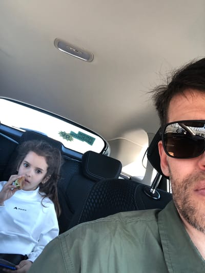This image captures a candid moment inside a car, likely a selfie or a photo taken by the adult subject, with a skewed, diagonal composition. The setting is a car interior during bright daylight, suggested by the light streaming through the windows.

The primary subjects are an adult male and a young girl. The adult male, partially visible in the upper left, has dark hair, stubble, and is wearing sunglasses that reflect the car's interior. He is dressed in an olive green or khaki-colored shirt or jacket. A metal rod, possibly part of a headrest mechanism, is visible near his shoulder.

In the lower right, a young girl with long, dark brown hair is seated in the back seat. She wears a white long-sleeved shirt featuring a black triangular logo with the word "PULKA" beneath it. She holds a small, light brown food item, possibly a pastry or bun, in her right hand, and looks directly at the camera with a direct and curious expression.

The car's interior features a light gray headliner with a rectangular overhead light fixture. The car seats are dark gray or black with a textured pattern. Through the rear window, bright outdoor light is visible, and a green and yellow sticker is affixed to the glass. Text on the sticker appears partially reversed and upside down, showing "NUSA," "WONGNUP," and "90 %" with a star shape. The scene depicts a casual, everyday activity, likely driving or resting in a car, in what is identified as Lisy Nos, Russia.