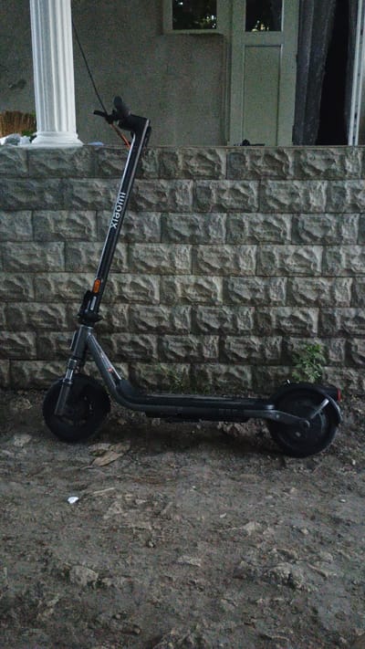 The image shows an electric scooter leaning against a textured stone wall. The scooter is black and has the "xiaomi" logo visible on its stem. It is positioned horizontally across the frame, with its front wheel towards the left and its back wheel towards the right. The scooter's handlebars, stem, and frame are clearly visible.
The background consists of a wall made of irregularly shaped, light-colored stones arranged in a stacked pattern. To the left of the wall, a portion of a building is visible, featuring a window and a white column. The ground beneath the scooter appears to be rough, earthy terrain with some scattered debris.
There is no discernible activity taking place; the scooter is stationary. The lighting suggests it is daytime, but the overall mood is somewhat muted, perhaps due to overcast weather or low light conditions. There are no people visible in the image. The location context provided is Mitoc, Moldova, but no specific landmarks within the image definitively confirm this. The text "xiaomi" is visible on the scooter.