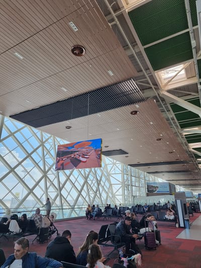 The image depicts the interior of a modern airport terminal, possibly in Mora, Northern Cyprus/Cyprus. A high, geometrically patterned glass roof allows ample sunlight to enter. The airport walls feature a layered construction with what appear to be ventilation panels. A large advertisement or digital display hangs suspended from the ceiling.

Rows of seated passengers occupy the foreground, many with luggage. Some are engaged in conversation or using electronic devices. A red-carpeted area runs alongside the seating. Further down the terminal, more passengers are visible, waiting near gates or information kiosks. The atmosphere suggests a busy airport environment.