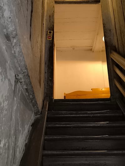 The image shows a dark, narrow staircase leading upwards. The walls on either side of the staircase are textured and appear aged, with one side being a rough, light-colored material and the other a darker, wooden panel. A small, yellow sign with red and black symbols is attached to the wooden panel near the top of the visible stairs. At the top of the staircase, an open doorway reveals a brightly lit room with a slanted, wooden ceiling and a plain white wall. A low, wooden headboard of a bed is visible against the white wall. The overall impression is of a rustic and somewhat aged interior space. This image was taken in Kuldīga, Latvia.