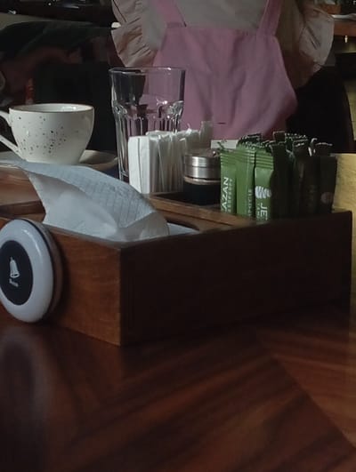 A table in a cafe or restaurant in Baktuu-Dolonotu, Kyrgyzstan. On the table is a white speckled teacup, a glass of water, a wooden box filled with napkins and sugar packets, and a white call button. A person wearing a pink apron is visible in the background. The table is made of dark wood.