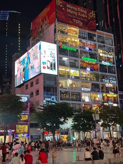 This is a nighttime, street-level photograph taken in Ho Chi Minh City, Vietnam. The scene is bustling with activity, featuring a multi-story building adorned with numerous brightly lit signs for various businesses, including "CƠM TẤM CALI," "AMCALI," "KANGAROO," "THE LETTER," "1971's CAFE," "Michi. Sushi," "THE ONE," "DOWNTOWN," and "LAST PIZZA." Some of the signs are in Vietnamese, while others are in English.
In front of the building, a large red banner with Vietnamese text that translates to "Happy New Year, Happy Spring, Land of Vietnam Renewed" is prominently displayed. Sidewalks are filled with pedestrians, many of whom appear to be casually walking or gathering. Some are captured in motion, suggesting movement and social interaction. The lighting is artificial, coming from the signs, streetlights, and what appear to be illuminated windows of the building. The overall atmosphere seems lively and energetic, typical of a busy urban environment at night. There are also trees and street furniture visible, adding to the urban streetscape. The image is vertically oriented, with the top of the frame appearing on the left side.