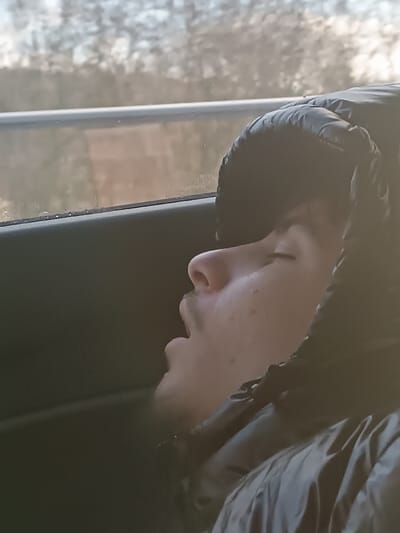 The scene is located in Stadtkyll, Germany. The image features a male individual, appearing to be a young adult, positioned in profile inside a vehicle. His eyes are closed and his mouth is slightly ajar, indicating a state of sleep or repose. He has light facial hair on his chin and upper lip, and minor dermatological blemishes are visible on his cheek. He wears a black, padded, hooded jacket with the hood covering his head. The background, viewed through a vehicle window, is blurred and depicts an outdoor environment with indistinct vegetation and a horizontal light-colored barrier. Specific landmarks to confirm the precise location within Stadtkyll are not discernible.