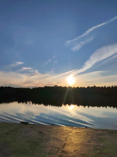 This vertical, full-frame photograph captures a serene sunset over a body of water, likely a lake or river, in Nifantovo, Russia. The scene is dominated by a vast, clear blue sky transitioning to warmer hues of orange and pink near the horizon. Wispy white clouds streak across the sky, some shaped like contrails, adding texture and depth. The sun, a bright, almost blinding orb, is setting just above a dark silhouette of a forest line that borders the water. Sunbeams radiate outwards from the sun, creating a lens flare effect and casting a warm glow.
The water's surface is calm, reflecting the sky and the distant treeline with remarkable clarity. Gentle ripples, possibly from a passing boat or the breeze, create a mesmerizing pattern of light and shadow on the water. The foreground consists of a sandy or earthy shoreline, with a subtle slope leading down to the water's edge. There are a couple of dark, indistinct objects on the shoreline, possibly mooring posts or small pieces of equipment. A faint, pale rainbow or atmospheric halo is visible in the sky above the treeline, to the left of the sun.
The time of day is clearly sunset, indicated by the low angle of the sun and the warm colors in the sky and water's reflection. The weather appears clear and pleasant, conducive to enjoying the outdoors. The presence of the forest line and the vast sky suggests a natural, possibly rural or semi-rural, setting. The overall mood of the image is peaceful and tranquil, evoking a sense of calm and natural beauty. There is no visible text in the image.
