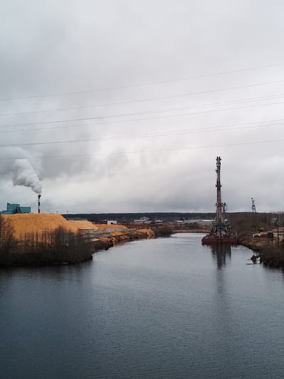 The image depicts a river with industrial elements on both banks under an overcast sky. On the left bank, a large pile of wood chips or sawdust is visible, alongside a factory or industrial building with a prominent chimney emitting a plume of smoke. On the right bank, there are two large cranes, one on a barge in the river. Power lines stretch across the sky. The scene suggests an industrial area, possibly a lumber mill or related facility, along a waterway. The location of Depo, Russia, is not confirmable with this image alone.