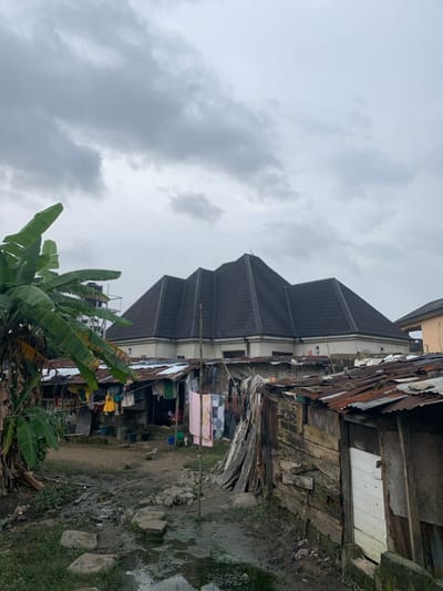 A large building with a dark gray roof stands next to weathered structures made of wood and corrugated metal. Laundry hangs on a line strung between buildings. Banana trees grow in the foreground. The ground is muddy and uneven. A cloudy sky fills the background, suggesting overcast weather. The scene depicts a stark contrast between the newer building and the older, more dilapidated surroundings.