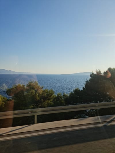 The image captures a scenic view of the Adriatic Sea from a coastal road in Drvenik, Croatia. The sky is a clear, bright blue, with a gentle gradient towards the horizon. The sea stretches out, a deep blue expanse dotted with small ripples. In the distance, the soft outlines of islands or peninsulas are visible against the horizon, their forms slightly blurred by atmospheric haze.
In the foreground, a dense line of green trees borders the coastline, with some trees appearing closer and more detailed, showcasing their leafy canopies. A metallic guardrail runs along the edge of the road, casting a long shadow across the asphalt. The road itself is paved and appears to be a typical coastal highway. Reflections are visible on the surface of what is presumed to be a window, suggesting the photograph was taken from a vehicle. The overall impression is one of a sunny day, likely in the late afternoon given the long shadows, offering a tranquil and picturesque view of the Croatian coast.