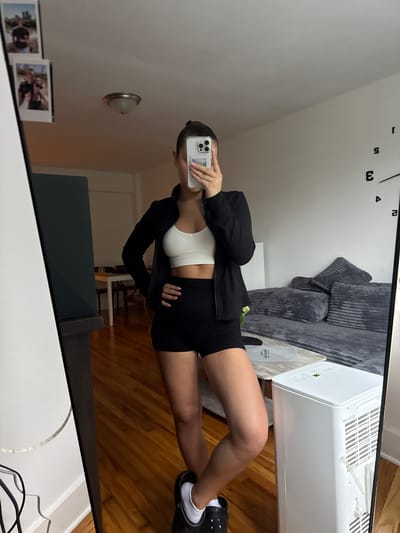 A young woman stands in front of a large mirror, taking a selfie with her phone. She is wearing a white sports bra, black shorts, and a black zip-up jacket. Her hair is pulled back in a bun. The mirror reflects a room with wooden floors, a couch, and a portable air conditioner. In the background, a dining table and chairs are visible. The room appears to be an apartment in the City of Yonkers, United States of America.